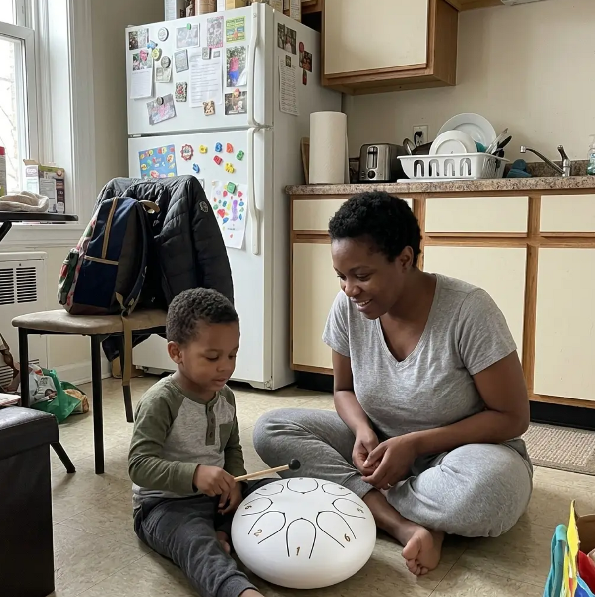 A boy tapping a white drum on the kitchen floor while his mother watches